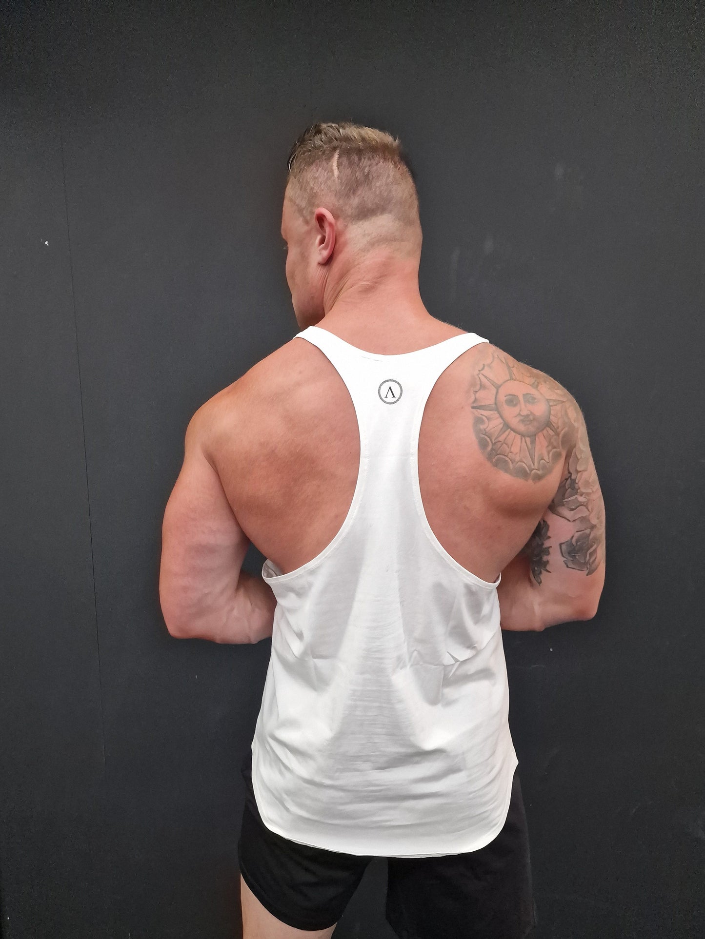 Man in a white tank top and black shorts lying on a dark surface with tattoos visible.