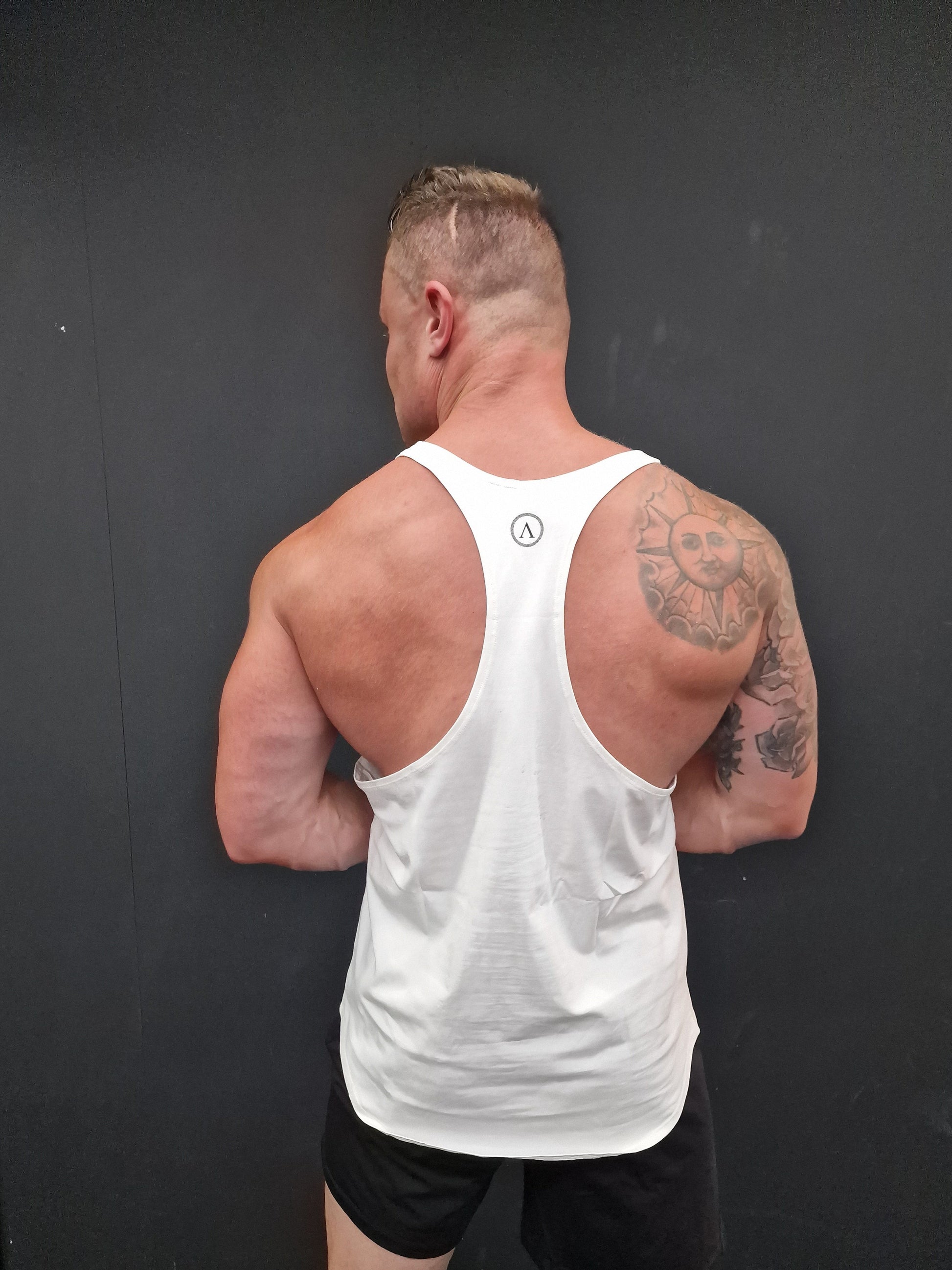 Man in a white tank top and black shorts lying on a dark surface with tattoos visible.